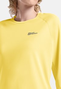 Gelbes Langarmshirt aus glattem Stoff; mit einem runden Ausschnitt und grauem Logo auf der linken Brust, sowie Raglanärmel für Komfort.