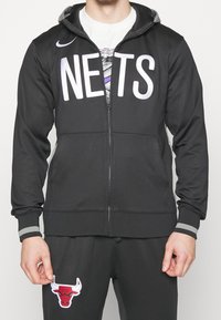 Veste zippée noire avec "NETS" en grandes lettres blanches. Accentuée de poignets gris et d'une capuche, associée à un pantalon de sport gris affichant un logo de taureau rouge.