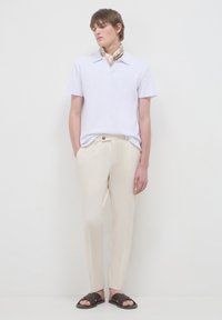 Polo shirt bianco a maniche corte, pantaloni beige su misura, sandali intrecciati marroni, sciarpa leggera attorno al collo, in piedi contro uno sfondo neutro.