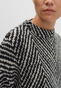Nahaufnahme einer Person, die einen schwarz-weißen, im Zickzack gestrickten Pullover mit hohem Kragen trägt. Ihr unteres Gesicht und der Hals sind zu sehen.