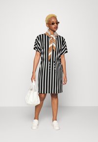 Vestido curto de manga curta com riscas preto e branco, com botões à frente, cintura atada, combinado com um lenço estampado, bolsa branca e sapatos.