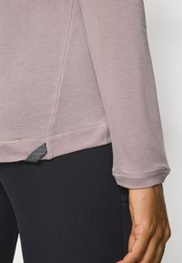 Langärmliges Oberteil aus hellrosa Stoff mit strukturiertem Muster, mit abgerundetem Saum und grauem Akzent an der Seitennaht, kombiniert mit schwarzen Leggings.