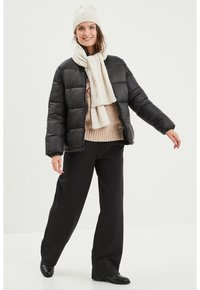 Giacca a puffo nera con chiusura a zip, abbinata a una sciarpa lavorata a maglia beige chiaro e un berretto. Il modello indossa pantaloni neri a gamba larga e stivali.