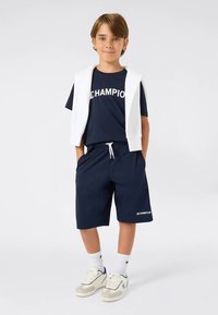 Zēns stāv ar rokām kabatās, ģērbies tumši zilās Champion šortos un t-kreklā, balts krekliņš uzmest uz pleciem un tennisbotes.