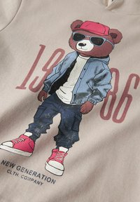 Ours portant une casquette rouge, des lunettes de soleil, une veste bleue, une chemise blanche, un pantalon foncé et des baskets rouges, avec le texte "1986" et "New Generation Cloth Company".
