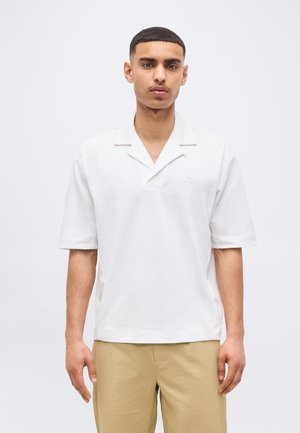 Junger Mann mit kurzen Haaren, der ein weißes kurzärmeliges Hemd mit Kragen und beige Hose trägt, steht vor einem schlichten hellen Hintergrund.