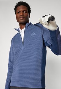Pullover sportivo blu con zip e coste testurizzate, con un colletto polo bianco sottostante. Un uomo tiene una mazza da golf indossando un guanto.