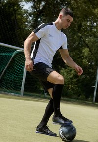 Sort Nike fodbold ved en spillers fødder. Spilleren har en hvid og sort kortærmet trøje, sorte shorts og knæhøje sorte sokker på.