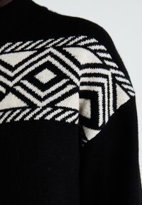 Svart tröja med ett geometriskt vitt mönster på den övre delen. Designen innehåller diamanter och zickzacklinjer i stickad textur.