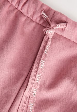Pantaloni della tuta in cotone rosa con una cintura elastica dotata di coulisse e dettagli bianchi con logo vicino alla cintura. Tessuto morbido e liscio.