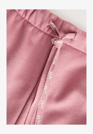 Pantaloni della tuta in cotone rosa con una cintura elastica dotata di coulisse e dettagli bianchi con logo vicino alla cintura. Tessuto morbido e liscio.