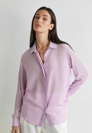 Femme aux cheveux longs et foncés portant un chemisier ample à boutons violet clair et un pantalon blanc, debout devant un fond uni clair.