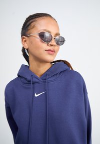 Sweatshirt azul-marinho com um logotipo branco da Nike, apresentando um capuz com cordão. O modelo usa óculos de sol ovais prateados com lentes escuras.