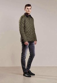 Man in een olijfgroene gewatteerde jas, grijze jeans en zwarte sneakers die tegen een effen beige achtergrond staat.