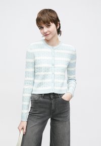 Svetlo modra črtasta jopica s teksturo kabelskega pletenja, okrogel izrez, zapenjanje na gumbe in dolgi rokavi, kombinirana s širokimi jeans hlačami v temno sivi barvi.