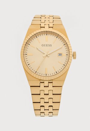 Montre Guess de ton doré avec bracelet métallique à maillons, cadran couleur champagne, lunette cannelée et guichet de date affichant le 20.