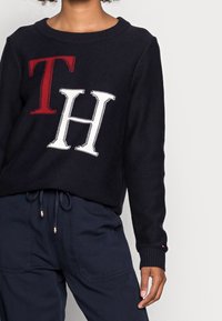 Granatowy sweter dziany z wyrazistymi czerwonymi literami 'T' i białymi 'H'. Okrągły dekolt, ściągacze na rękawach i u dołu. Połączony z ciemnymi spodniami.