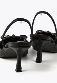 Svarta klacksandaler med en elegant design, försedd med tunna remmar, en slingrande detalj, och en smal, hög klack med kvadratisk bas. Guldhardwaredetalj.