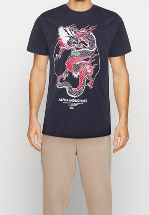 Mężczyzna w granatowej koszulce z czerwono-białą grafiką smoka i napisem „Alpha Industries”, w połączeniu z beżowymi spodniami, na neutralnym tle.