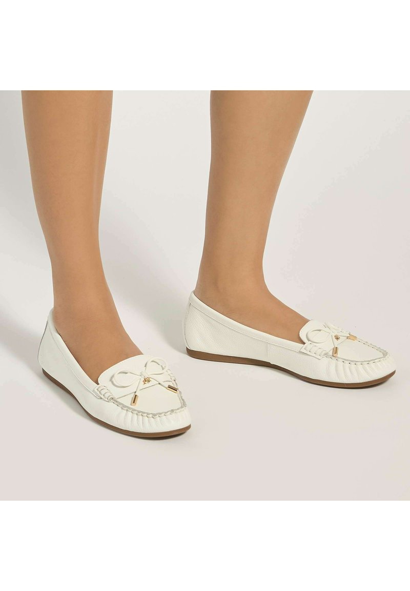 Mocassins en cuir blanc avec nœud décoratif, portés pieds nus, présentés sur un fond neutre clair.