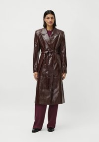 Donna che indossa un trench in pelle marrone scuro con cintura su pantaloni e camicia bordeaux, in piedi di fronte a uno sfondo bianco semplice.
