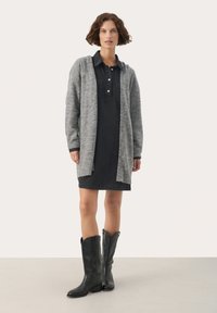 Cardigan tricotat gri peste o rochie din denim negru cu închidere prin butoane. Botine negre până la genunchi cu un design conic. Fundal simplu.