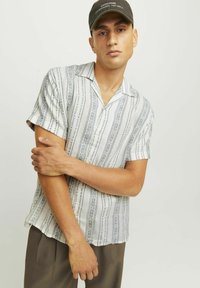 Chemise à manches courtes avec col, présentant une base gris clair ornée de motifs géométriques bleus. Associée à un pantalon marron ample.