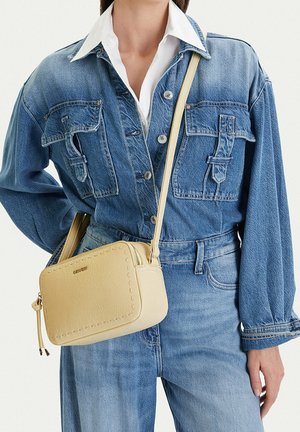 Mujer que lleva chaqueta y jeans de mezclilla azul, con un bolso cruzado pequeño de cuero beige con bordes cosidos.