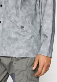 Jachetă texturată gri cu un buzunar cu buton, material neted și un model subtil, asociată cu pantaloni gri deschis cu detalii de coasere pe lateral.