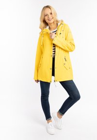 Giacca impermeabile gialla con cappuccio, caratterizzata da lacci bianchi, chiusura con zip e due tasche frontali. Indossata sopra una maglietta a righe e jeans blu.