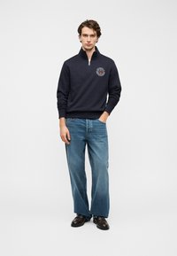 Πουλόβερ ναυτικού μπλε με ψηλό γιακά και φερμουάρ στο μπροστινό μέρος, με κυκλικό λογότυπο. Συνδυάζεται με ανοιχτό μπλε τζιν παντελόνι με φαρδιά πόδια και μαύρα παπούτσια.