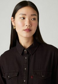 Chemise en denim noir avec des boutons-pression, deux poches sur la poitrine, un col et un petit emblème logo rouge près du bas. Texture lisse.