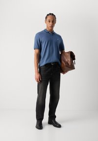 Tricou polo albastru cu mânecă scurtă, blugi închiși la culoare și pantofi negri. Ținând o geantă din piele maro. Design simplu, siluetă ajustată.