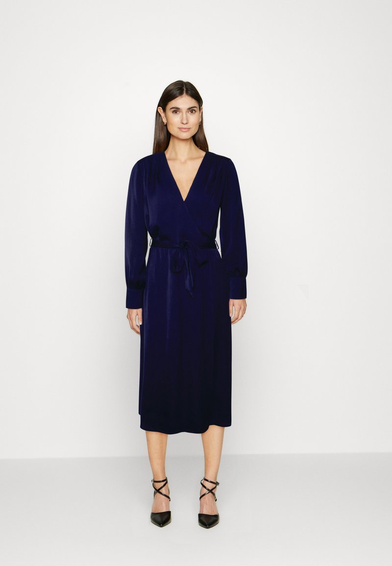 Vestido envelopado azul marinho com mangas longas, decote em V e fita na cintura. Textura suave e comprimento midi, combinado com saltos pretos de tiras.
