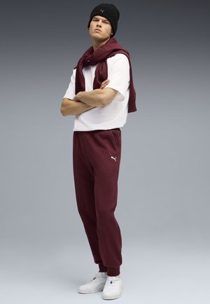 Bordeauxfarvede sweatpants med opkrøllede ben, parret med en hvid t-shirt. En matchende bordeauxfarvet trøje er draperet over skuldrene. Sort beanie-hat.