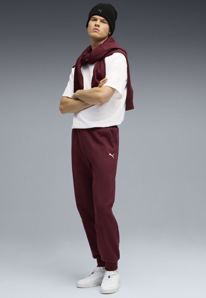Bordeauxkleurige sweatpants met omslagboorden, gecombineerd met een wit T-shirt. Een bijpassende bordeauxkleurige trui is over de schouders gedrapeerd. Zwarte beanie.