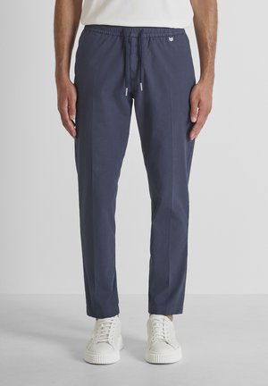 Man draagt donkerblauwe broek met trekkoord en witte sneakers, staand tegen een effen lichte achtergrond.