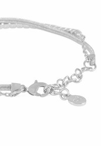 Silverfärgad armband med en blandning av kedjestilar, karbinhake-lås och en liten berlock med ingraverad logotyp. Slät, polerad yta.