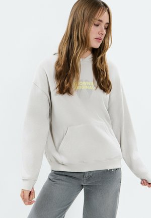 Joven con cabello largo que lleva una sudadera con capucha gris claro con texto amarillo y jeans grises, mirando hacia abajo a un lado contra un fondo blanco.