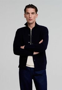 Cardigan lavorato a maglia blu navy con colletto alto e bottoni frontali. Indossato sopra una camicia color crema, abbinato a pantaloni in denim scuro. Tessuto morbido, design aderente.