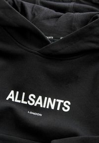 Sweat à capuche noir en tissu mélange de coton avec un logo blanc texturé "ALLSAINTS" et "LONDON" en dessous. Dispose d'une grande capuche et de poignets côtelés.