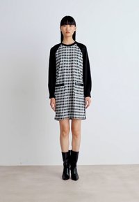 Mustavalkoinen houndstooth-kuvioinen mekko, jossa on mustat pitkät hihat, kaksi etutaskua ja erottuva trimmaus. Päällä polveen ulottuvat mustat saappaat.