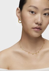 Pendientes de aro de oro con acentos texturizados y un delicado collar de oro con pequeños charms colgantes. Tonalidad de piel suave, blusa off-shoulder.