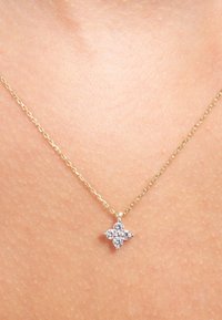 Gouden ketting met een klein, bloemvormig hangertje dat sprankelende heldere stenen bevat, rustend tegen de huid.