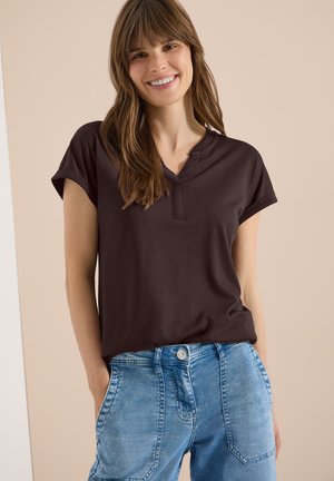 Femme souriante aux longs cheveux bruns portant un haut à manches courtes foncé et un jean en denim bleu clair, debout les mains dans les poches.