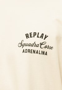 Ύφασμα κρεμ με μαύρο κείμενο "REPLAY Squadra Corse ADRENALINA" τυπωμένο σε τρεις γραμμές, η μεσαία σε πλάγια γραφή.