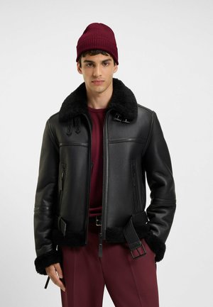 Schwarze Lederjacke mit einem plüschigen schwarzen Lammfellkragen und -manschetten, mit Reißverschlusstaschen und einem Schnallen-Detail in der Taille. Kombiniert mit einer bordeauxroten Mütze.