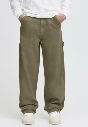 Persona che indossa pantaloni cargo verde oliva larghi, scarpe da ginnastica bianche e una maglia a maniche lunghe bianca con le mani nelle tasche.