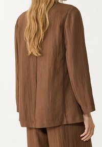 Femme aux longs cheveux blonds portant une veste marron texturée ample et un pantalon assorti, vue de dos.