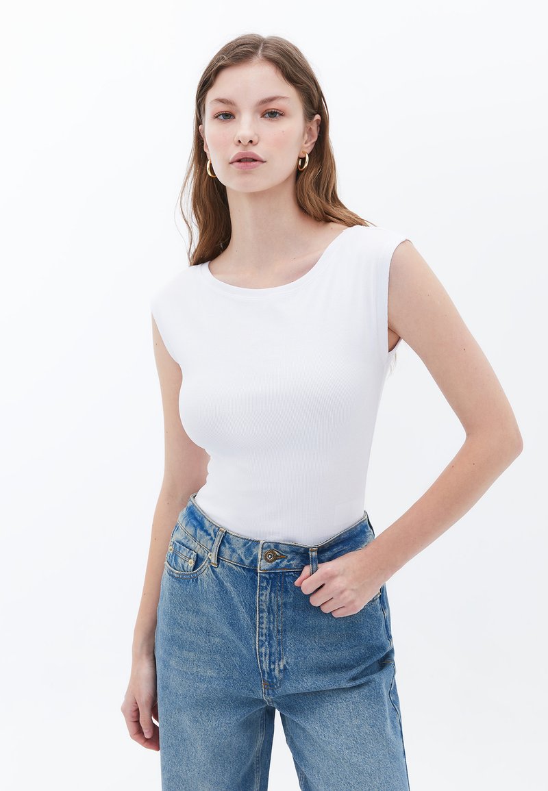 Witte geribbelde mouwloze top met een ronde halslijn, gecombineerd met hoge blauwe spijkerbroek met een knoop- en ritssluiting.